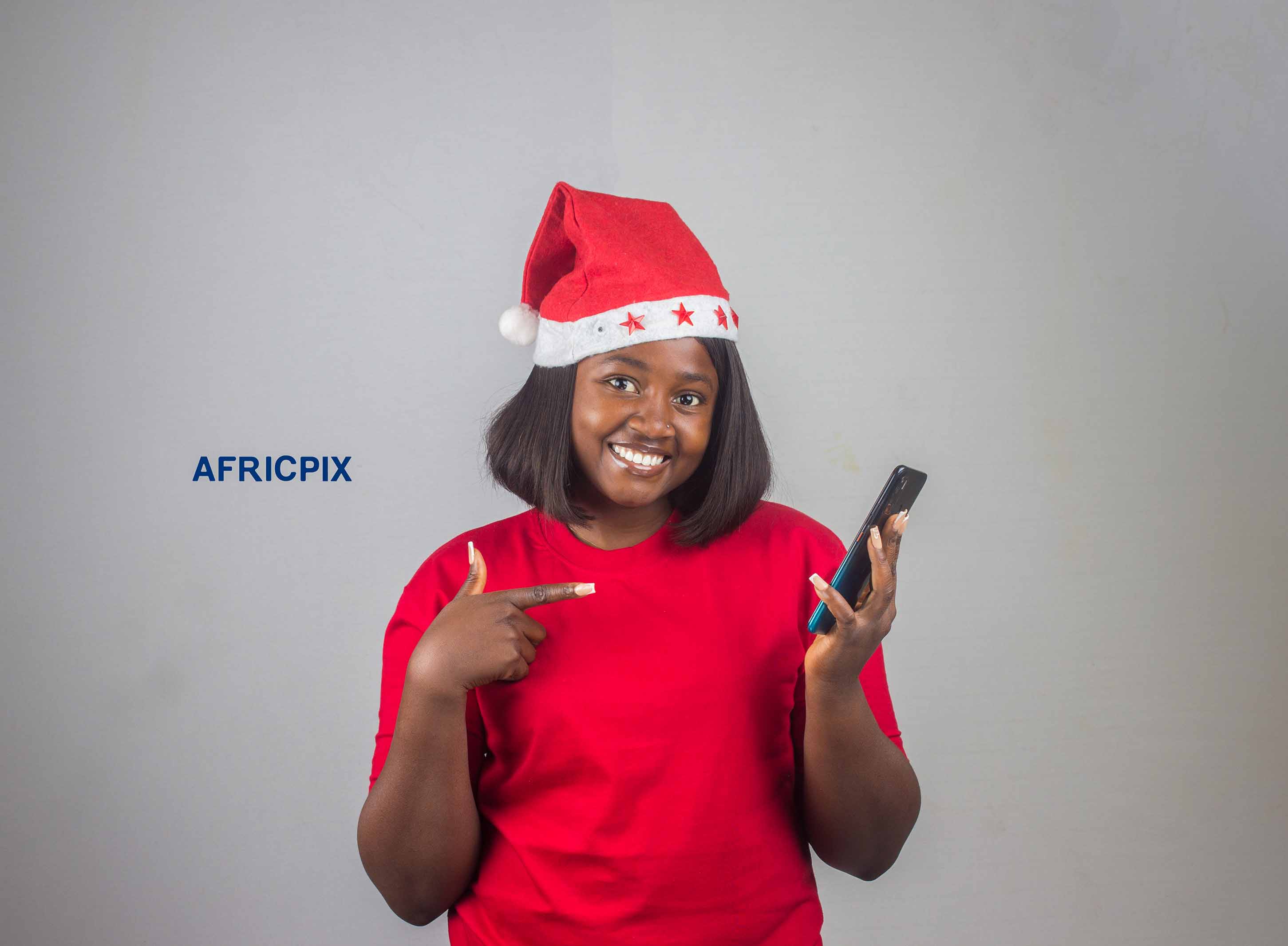 A happy Nigerian woman wearing a Christmas cap, holding a phone, and pointing at it with excitement.